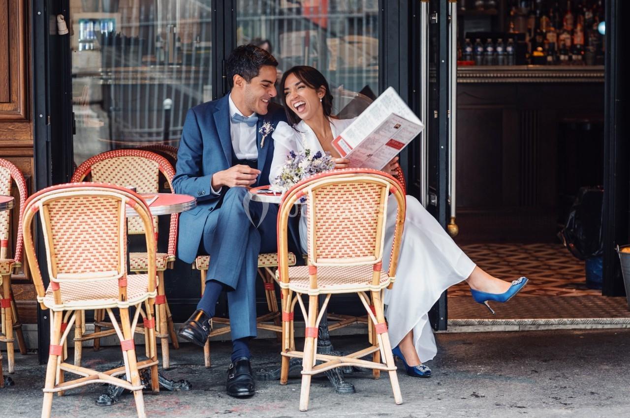 Un couple en tenue de mariage rit ensemble à une table de café en terrasse, dans un cadre urbain.
