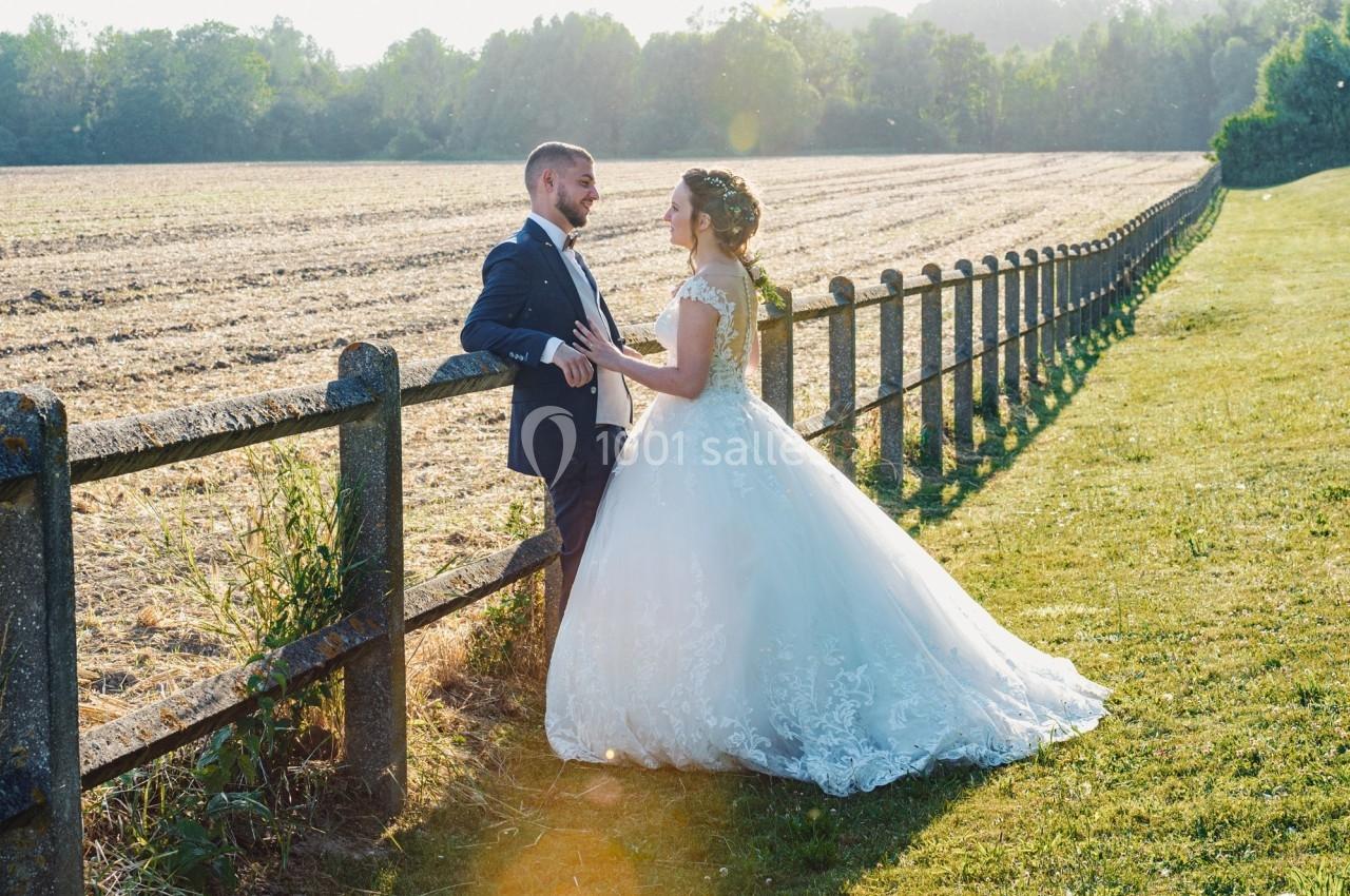 Un couple en tenue de mariage se tient près d'une clôture en bois, avec un champ et des arbres en arrière-plan.