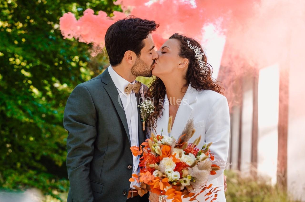 Un couple s'embrasse en extérieur, entouré de fumée rose, la mariée tenant un bouquet coloré.