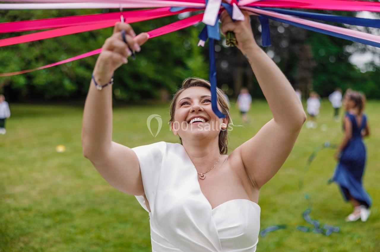 Une femme en robe blanche tisse des rubans colorés lors d'une activité en plein air sur une pelouse.