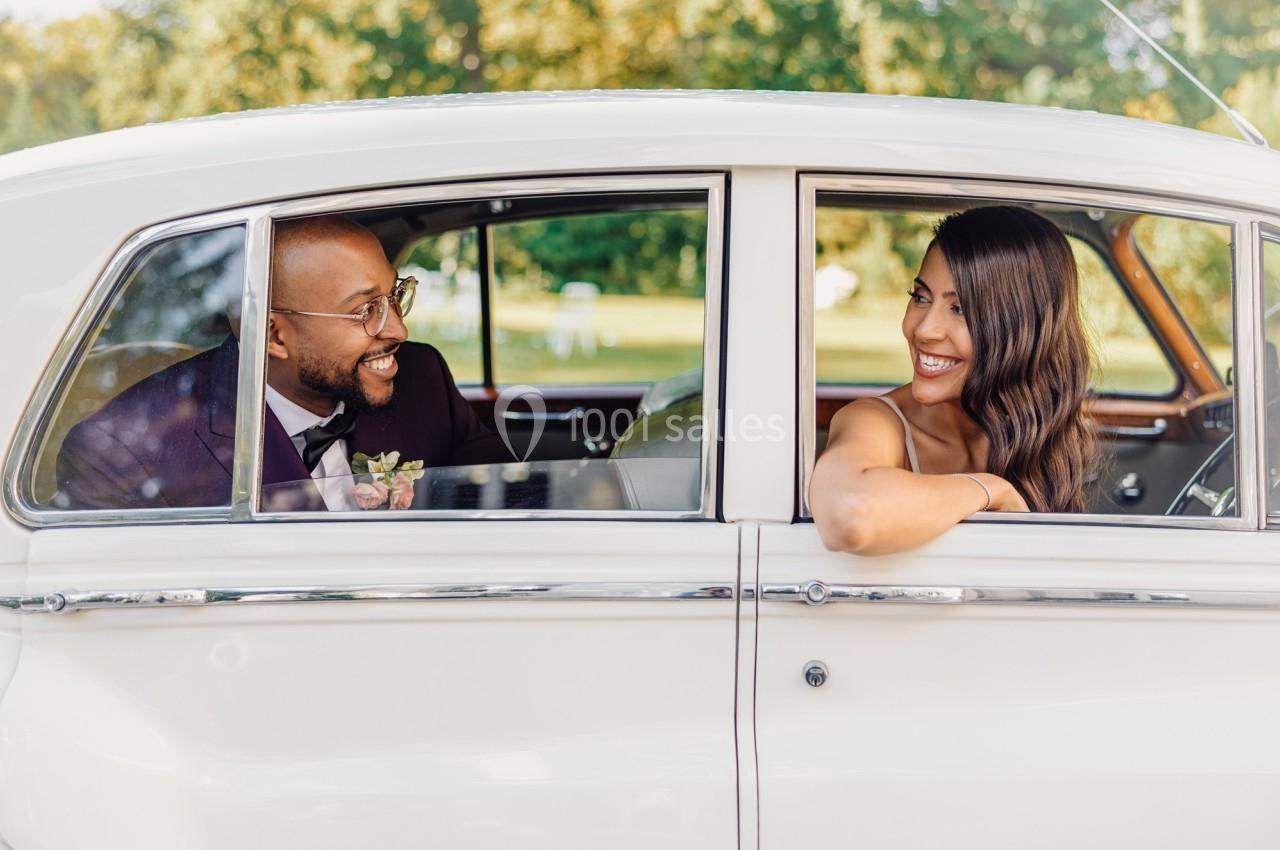 Un couple souriant assis dans une voiture ancienne blanche, avec un paysage verdoyant en arrière-plan.