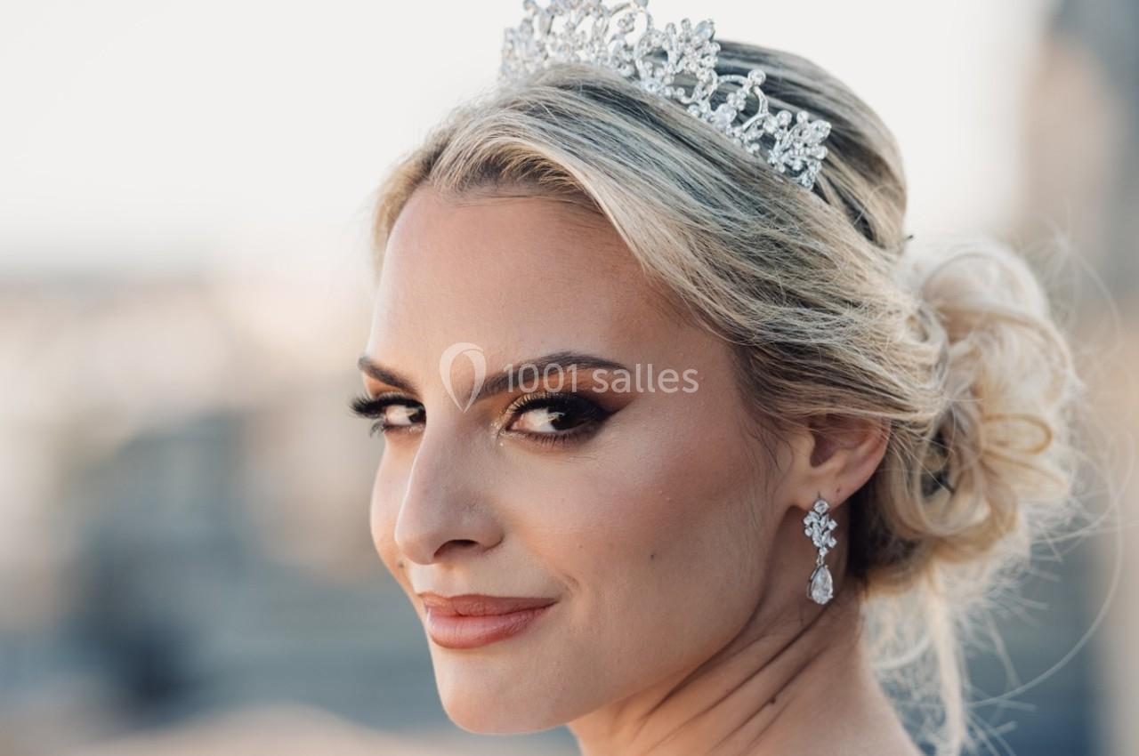 Femme blonde portant une couronne ornée de pierres et des boucles d'oreilles, coiffée d'un chignon élégant.