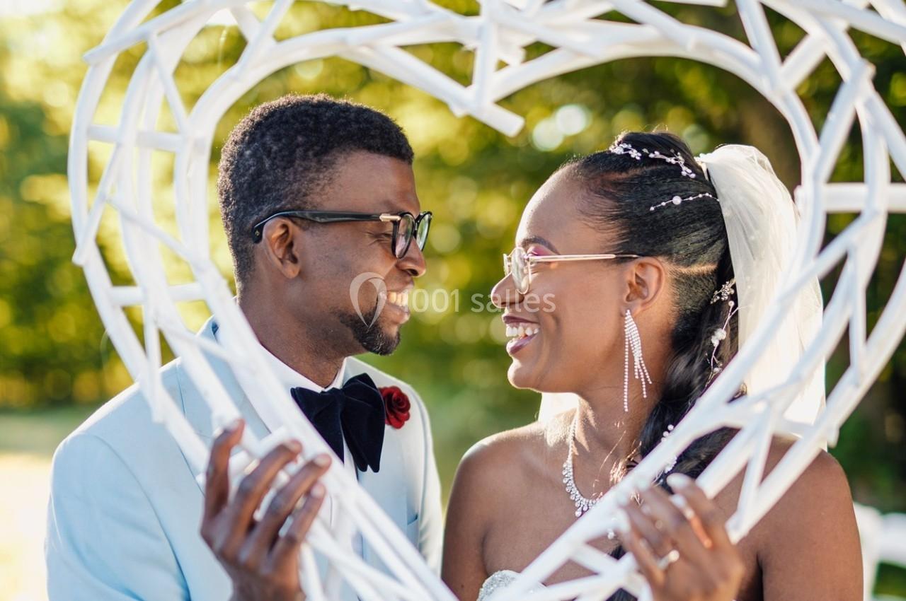 Un couple souriant se regarde à travers un cadre en forme de cœur, dans un décor extérieur ensoleillé.