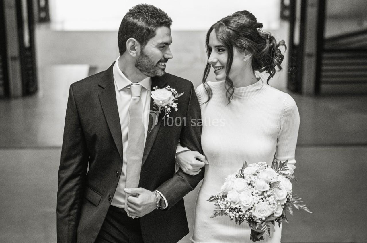 Un couple souriant en tenue de mariage marche bras dessus bras dessous, la mariée tenant un bouquet de fleurs.