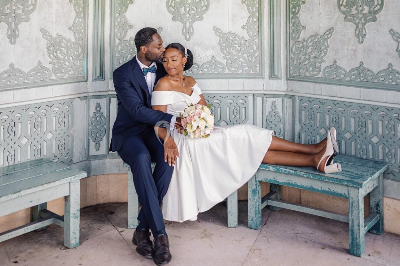 Un couple en tenue de mariage assis sur un banc, l'homme embrassant tendrement la femme qui tient un bouquet.