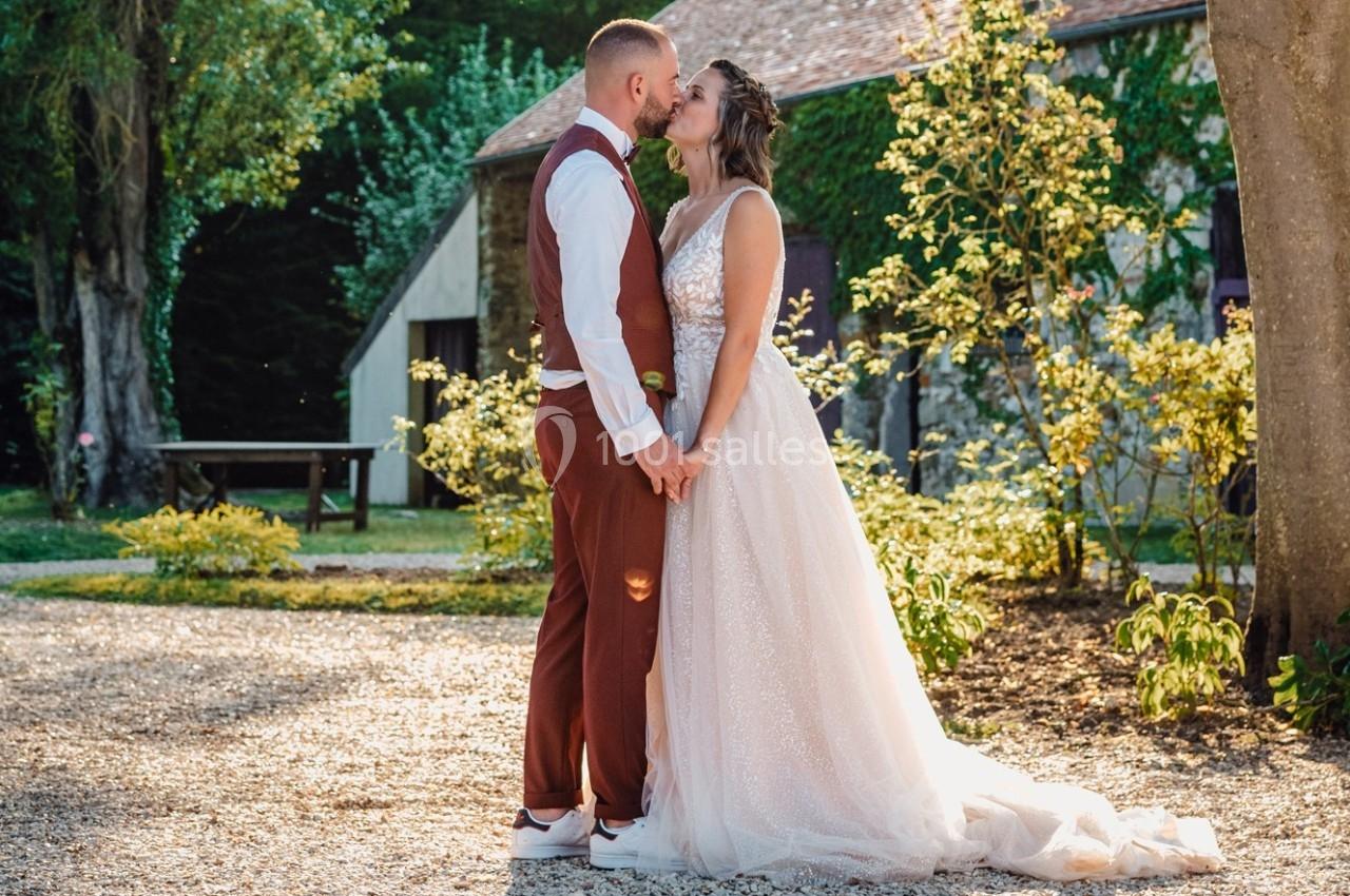 Un couple en tenue de mariage s'embrasse dans un jardin ensoleillé devant un bâtiment en pierre.