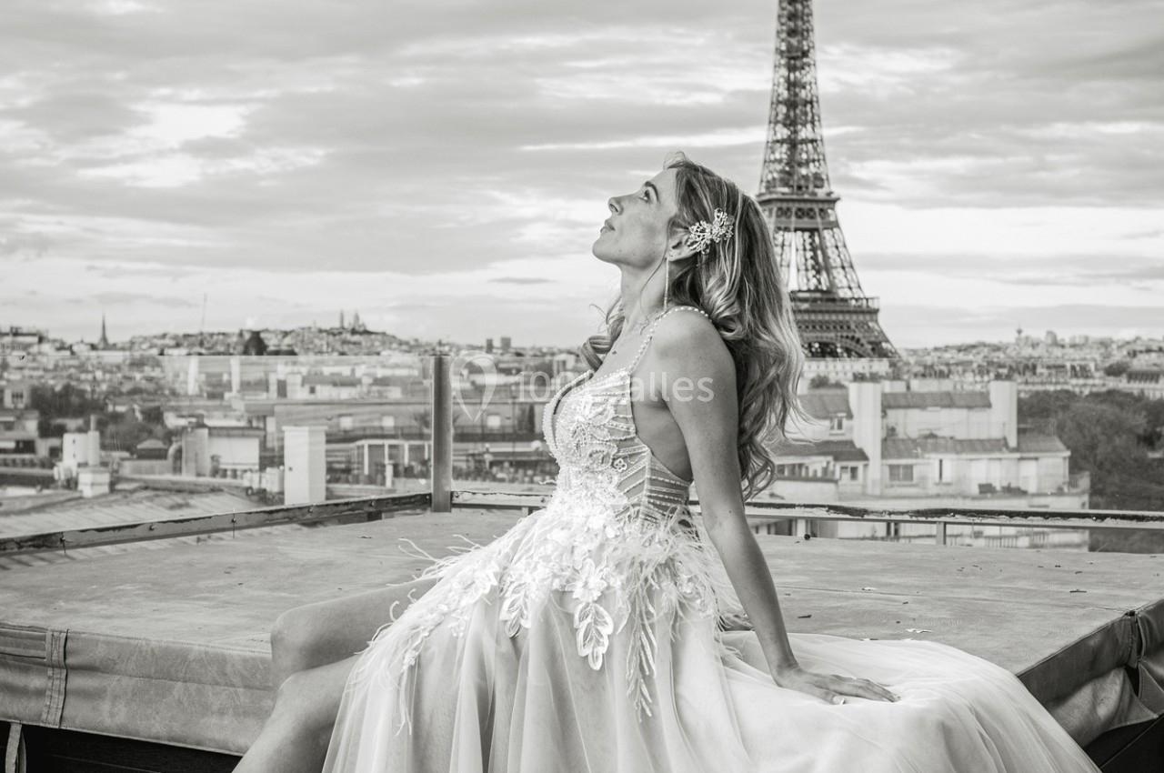 Femme en robe élégante assise sur un toit, avec la tour Eiffel et un ciel nuageux en arrière-plan.