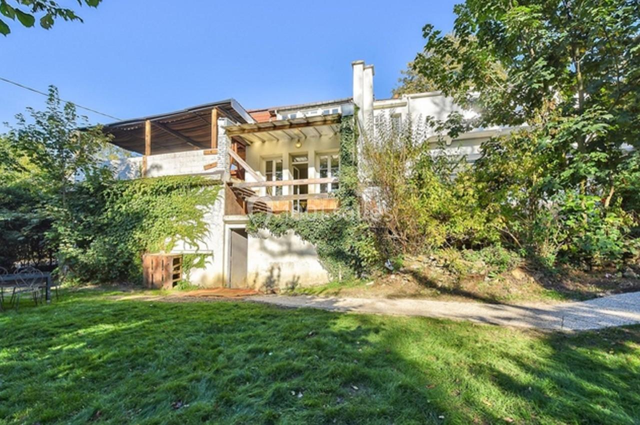Façade d'une maison entourée de verdure avec terrasse en bois et jardin arboré sous un ciel dégagé.