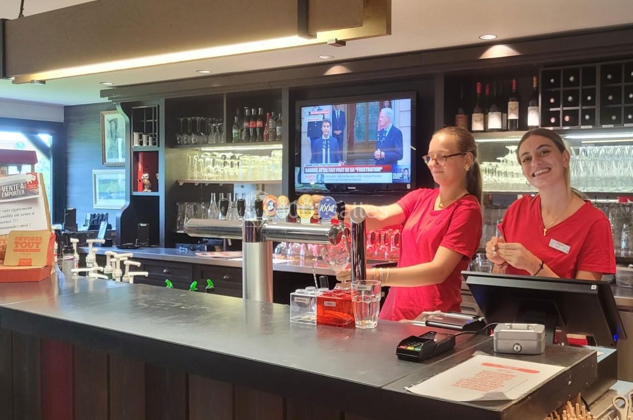 Deux employées en uniforme rouge travaillent derrière un comptoir de bar équipé de tireuses à bière et d'une caisse.