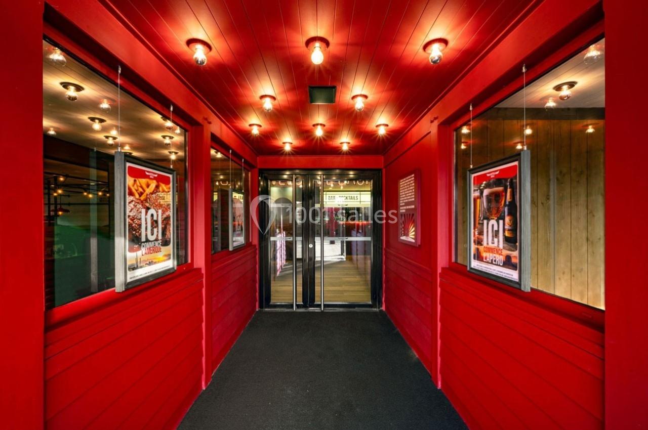 Entrée d'un bâtiment avec murs rouges, affiches publicitaires et portes vitrées sous un plafond éclairé.