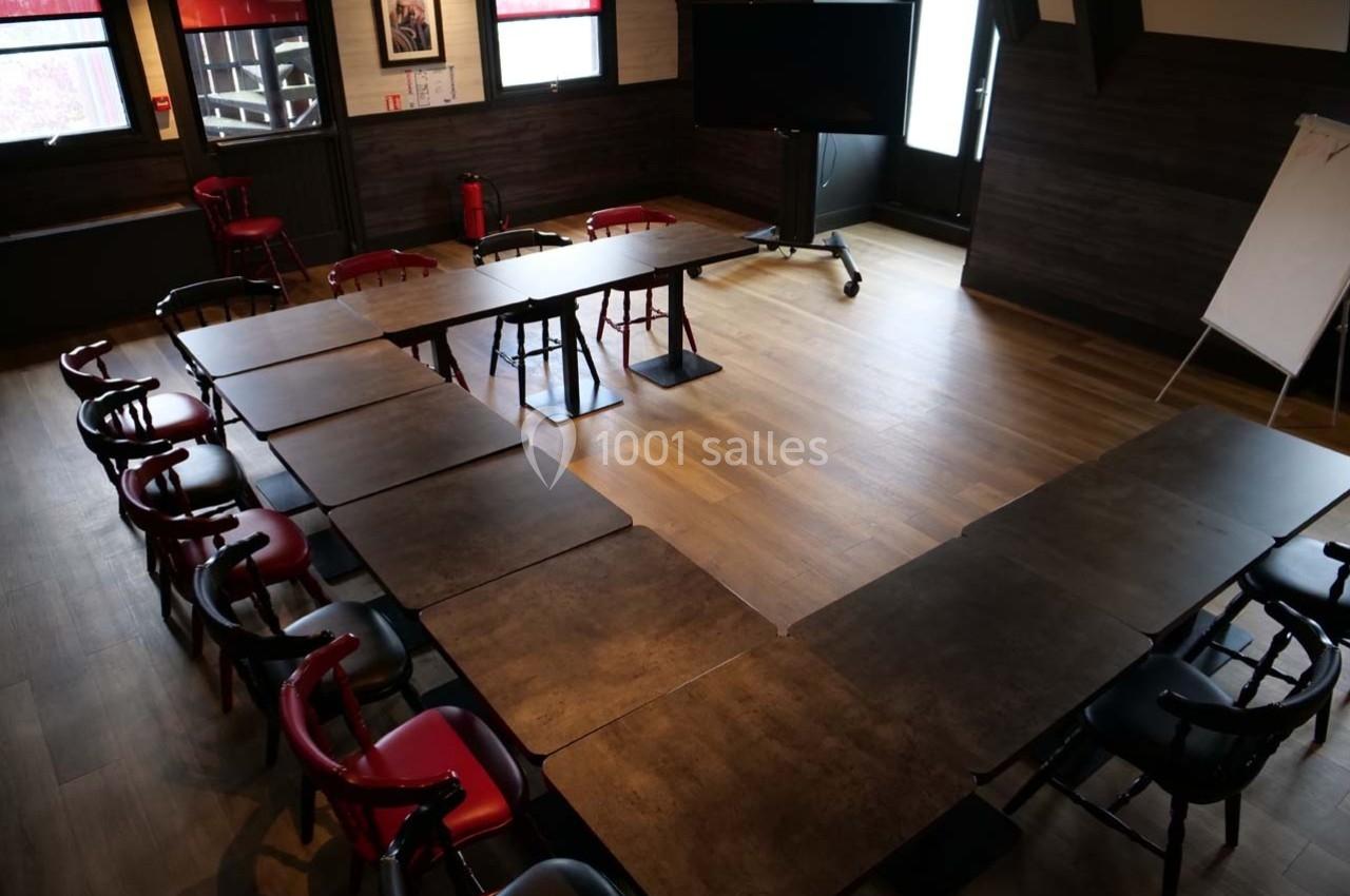 Salle de réunion avec tables disposées en U, chaises rouges et noires, tableau blanc et écran sur pied.