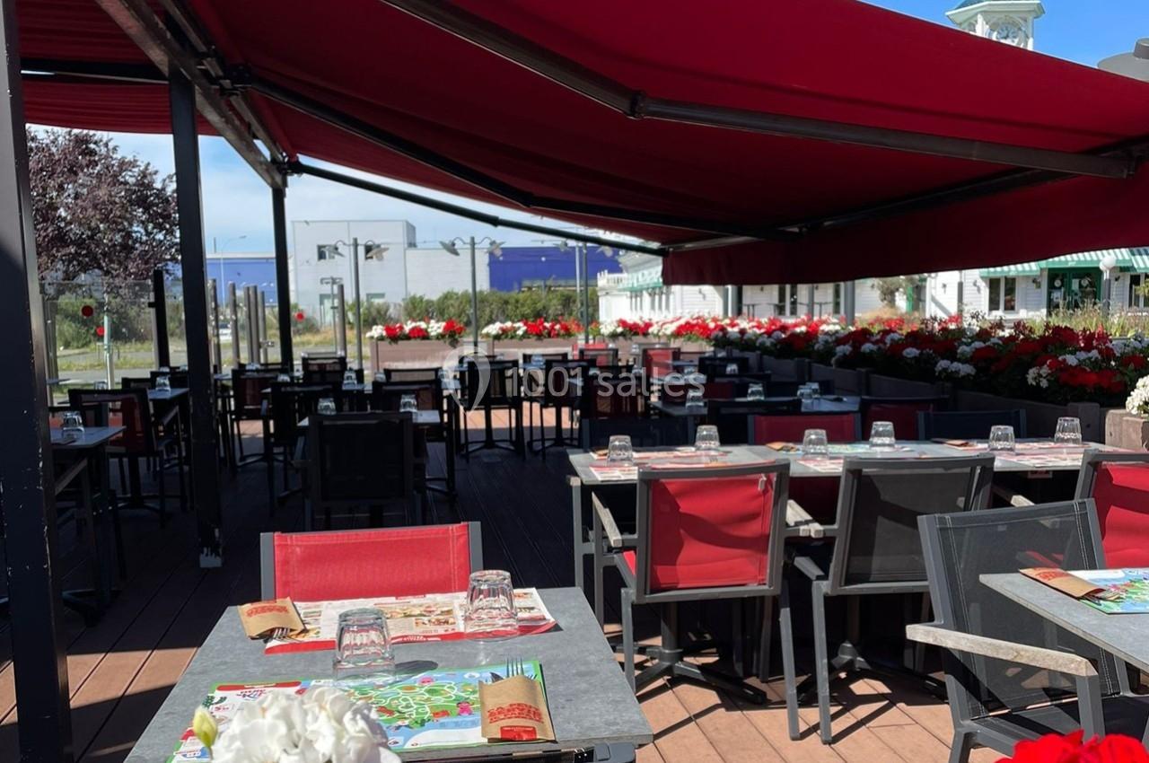 Terrasse d'un restaurant avec tables dressées, chaises rouges et noires, sous un grand auvent rouge, entourée de fleurs.