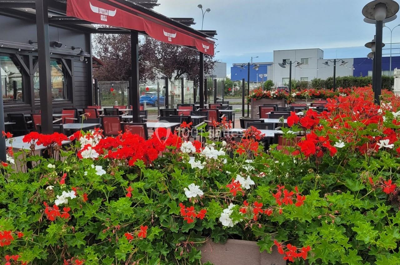 Terrasse de restaurant avec tables et chaises vides, entourée de jardinières fleuries de géraniums rouges et blancs.