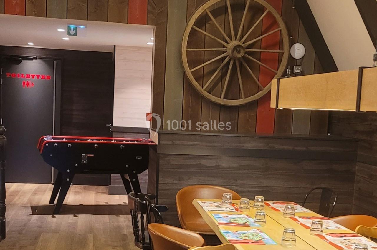 Salle de restaurant avec table dressée, baby-foot au fond et décoration murale incluant une roue en bois.