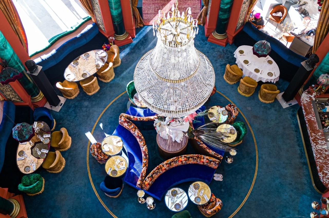 Vue plongeante d'une salle de restaurant luxueuse avec un grand lustre central, des tables rondes et une décoration colorée.