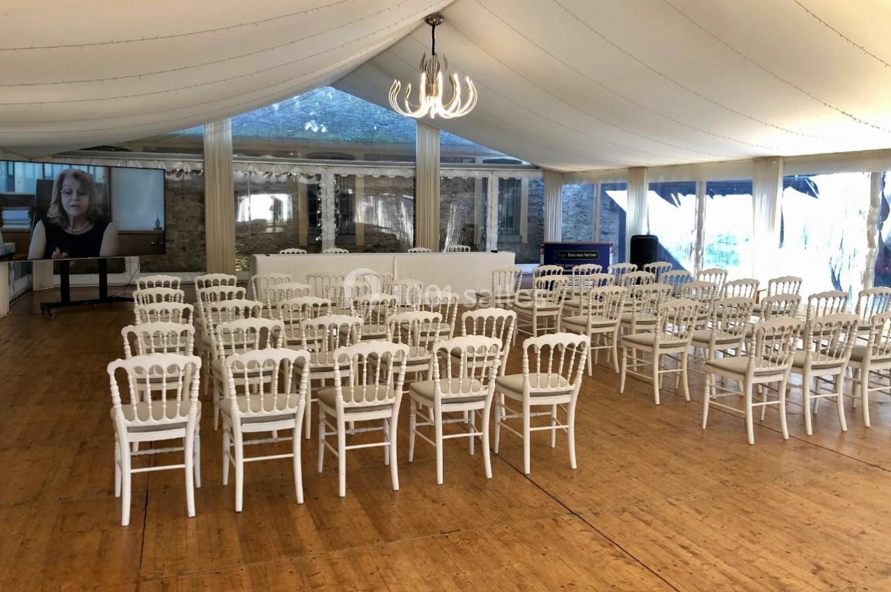 Salle de réception avec des chaises blanches alignées face à une table, sous une tente avec un lustre central.