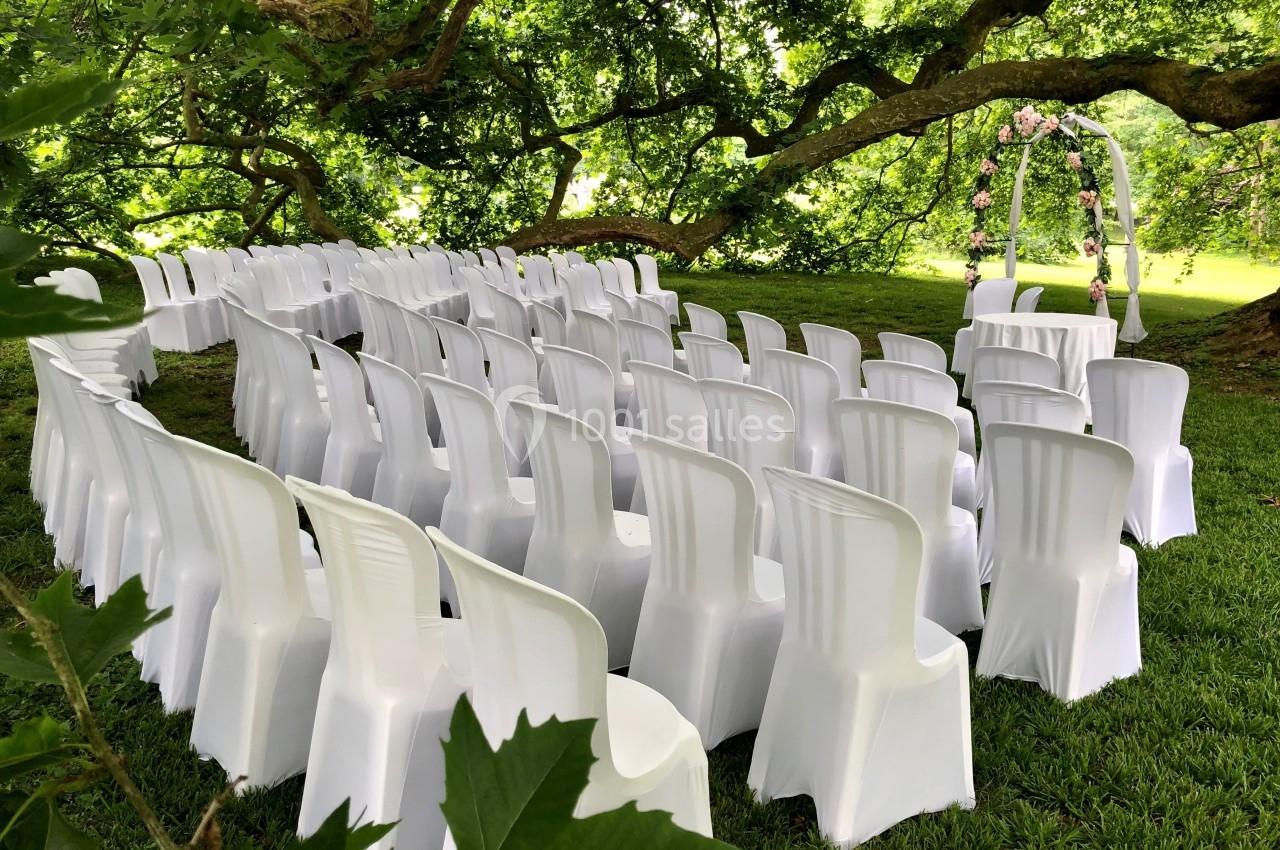 Chaises blanches disposées en rangées sous un grand arbre pour une cérémonie en plein air avec une arche décorée de fleurs.