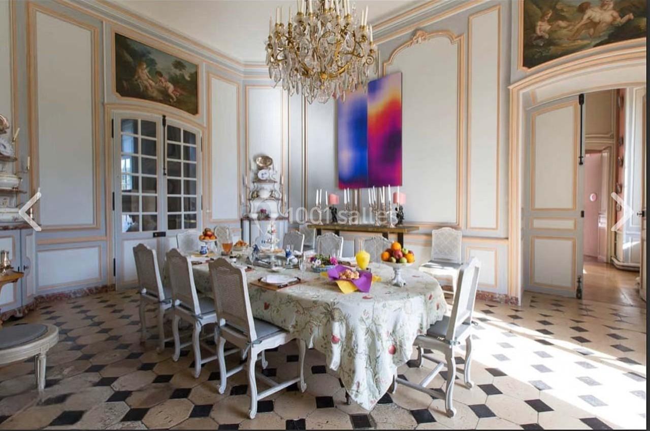 Salle à manger lumineuse avec table dressée, lustre en cristal, murs ornés et sol en carreaux noirs et blancs.