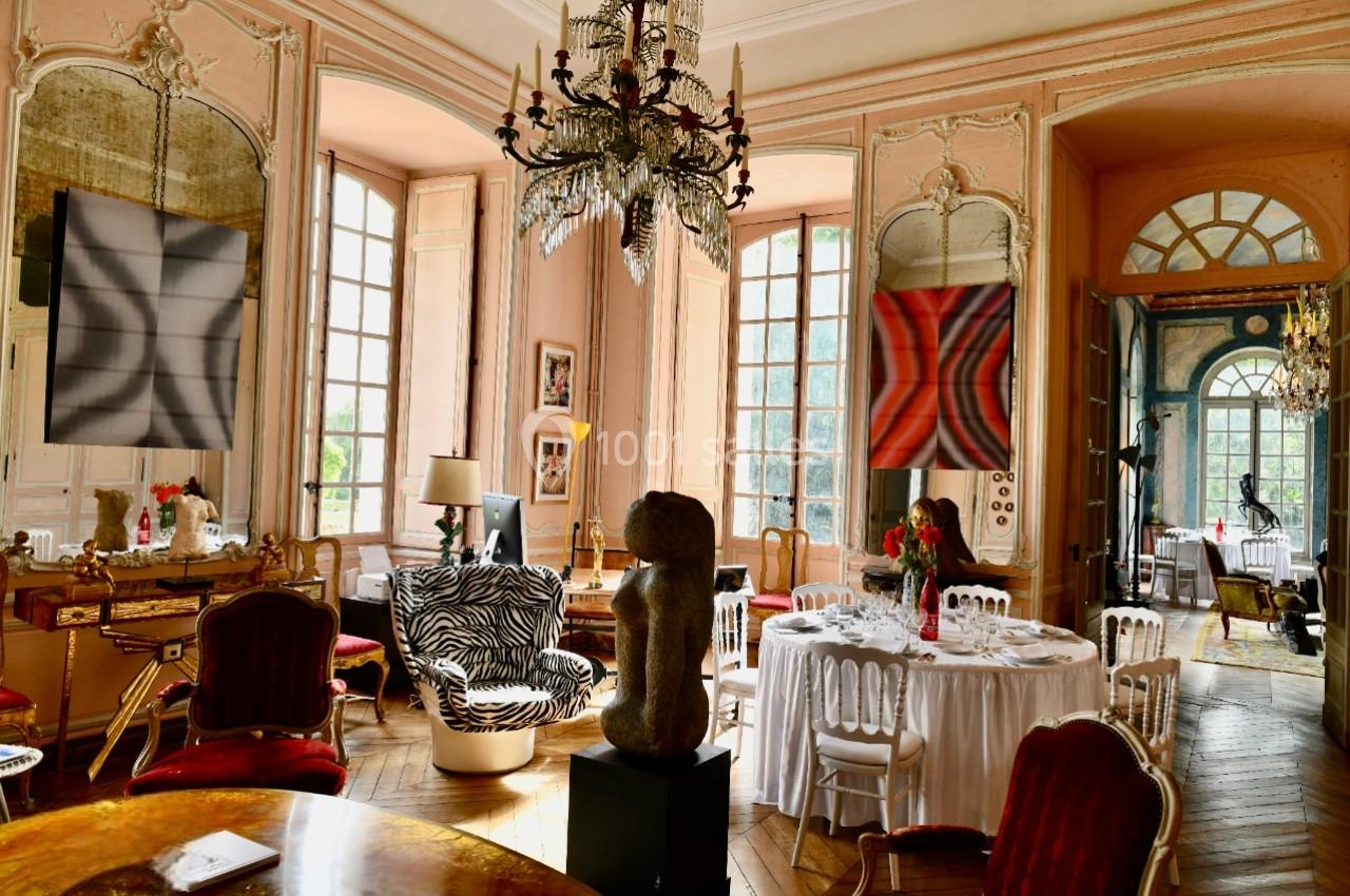 Salle élégante avec murs roses, lustre en cristal, mobilier ancien et tables dressées près de grandes fenêtres.