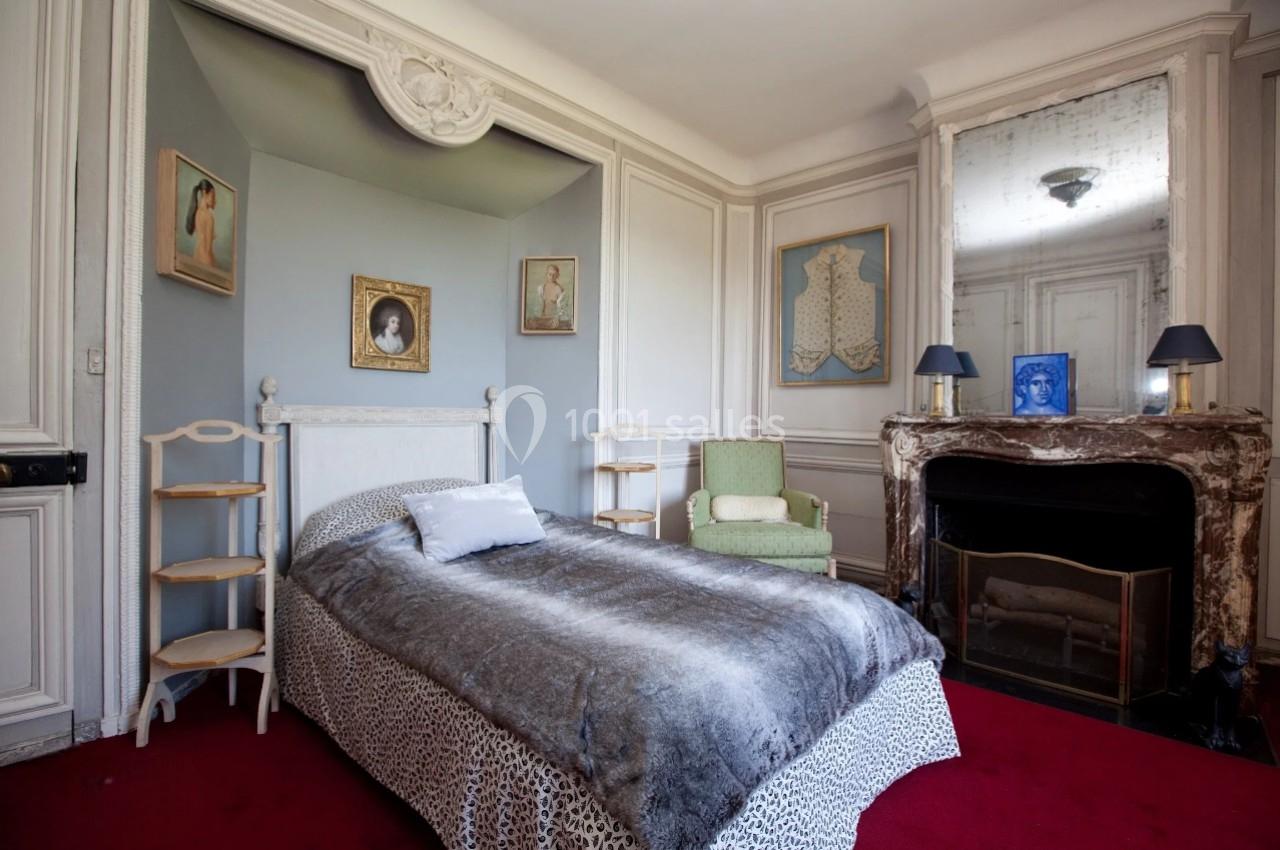 Chambre avec lit simple, cheminée en marbre, fauteuil vert, murs ornés de moulures et tableaux.