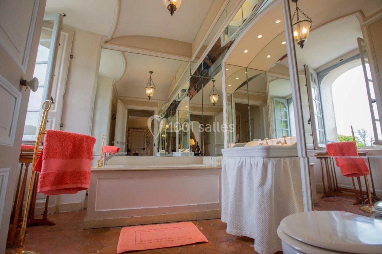 Salle de bain lumineuse avec miroirs, baignoire, serviettes orange et sol en carrelage rouge.