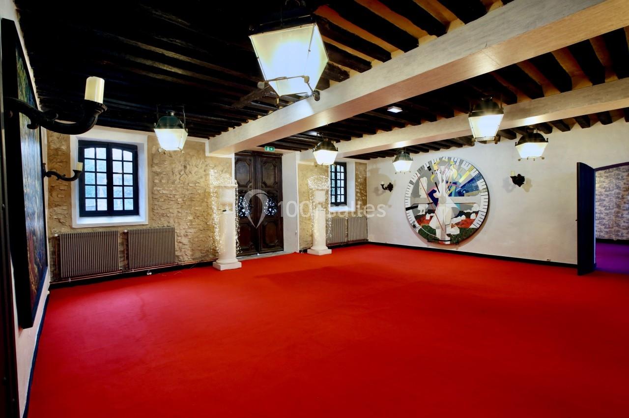 Salle avec murs en pierre, plafond à poutres apparentes, tapis rouge, luminaires suspendus et grande fresque murale colorée.