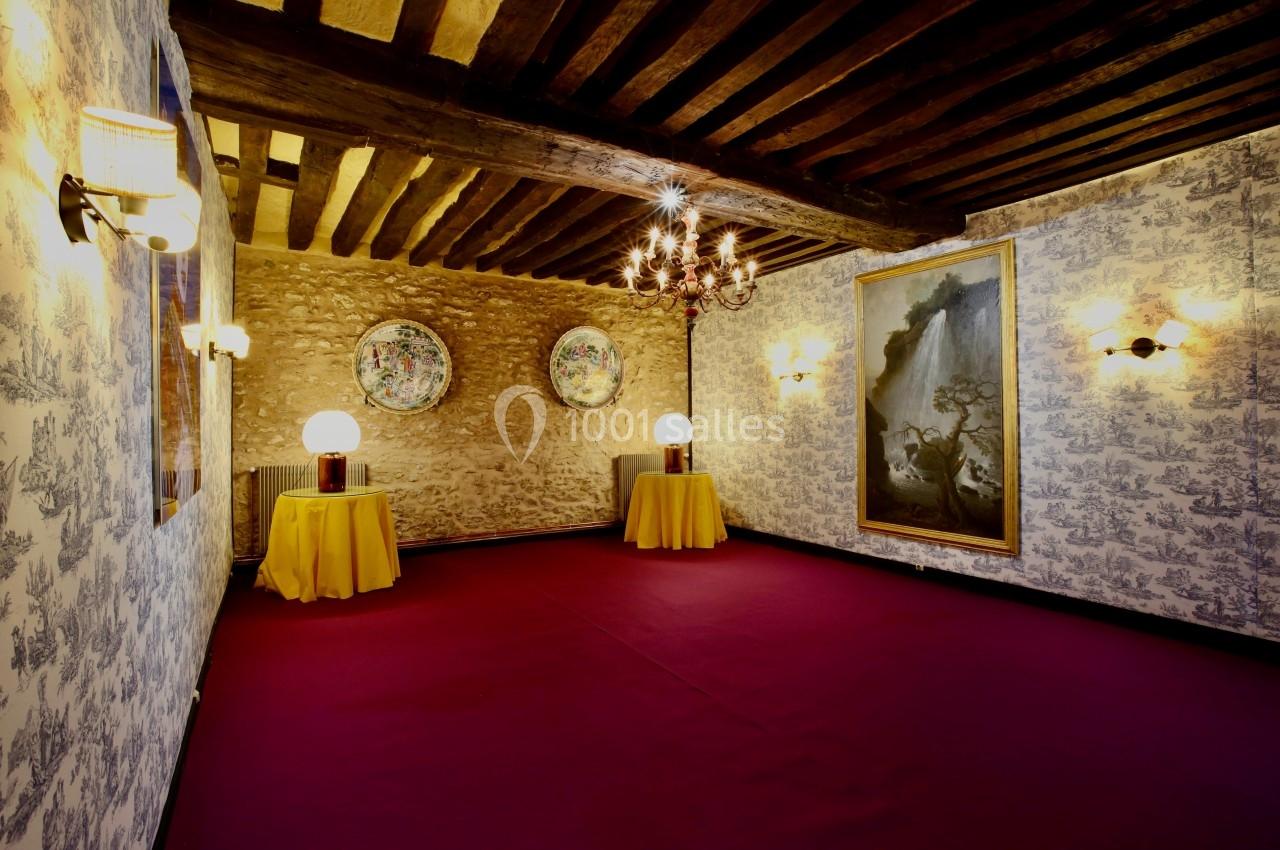 Salle avec poutres apparentes, murs en pierre et papier peint, éclairée par des lampes et décorée de tableaux et assiettes.