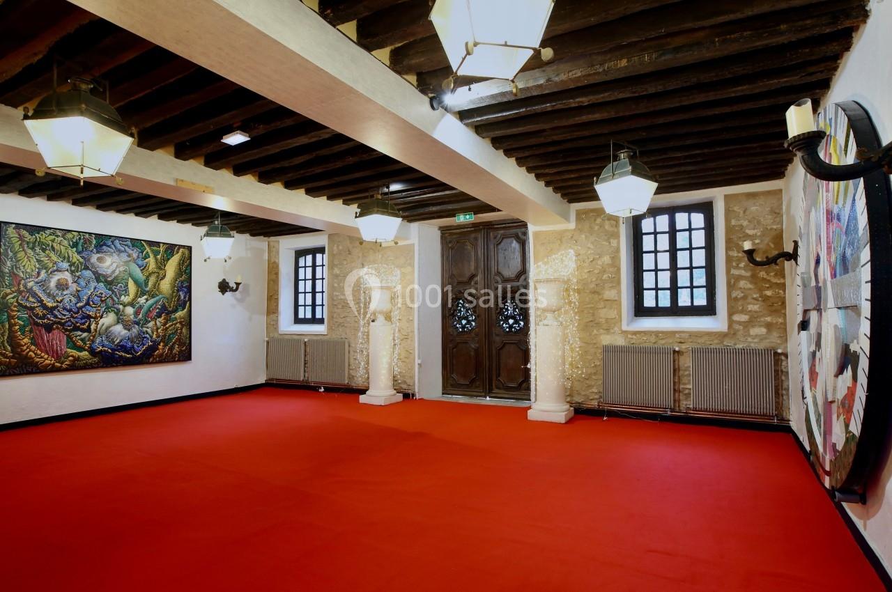 Salle avec un sol rouge, murs en pierre, poutres apparentes, œuvres d'art accrochées et colonnes décoratives.