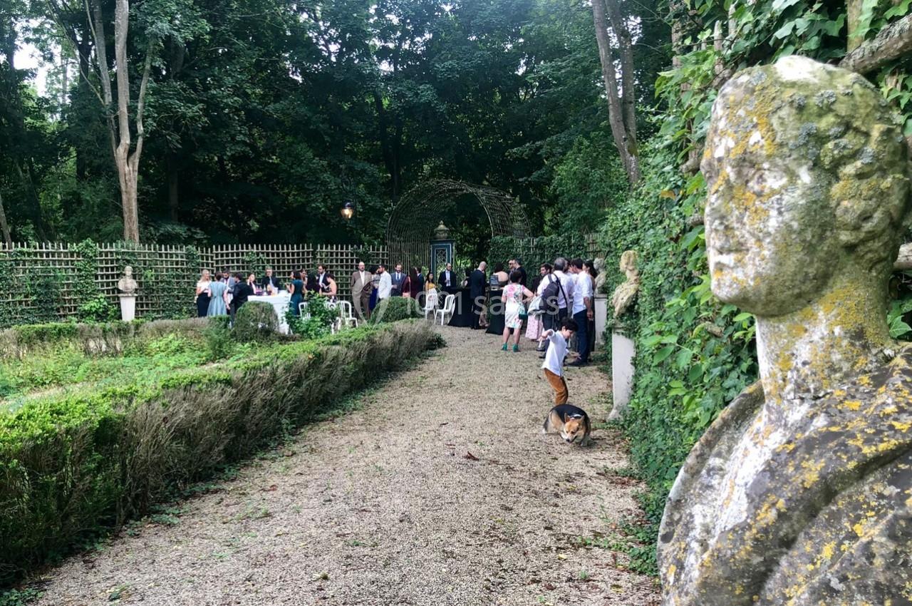Statue en pierre dans un jardin verdoyant où des personnes sont rassemblées près de tables et d'une arche.