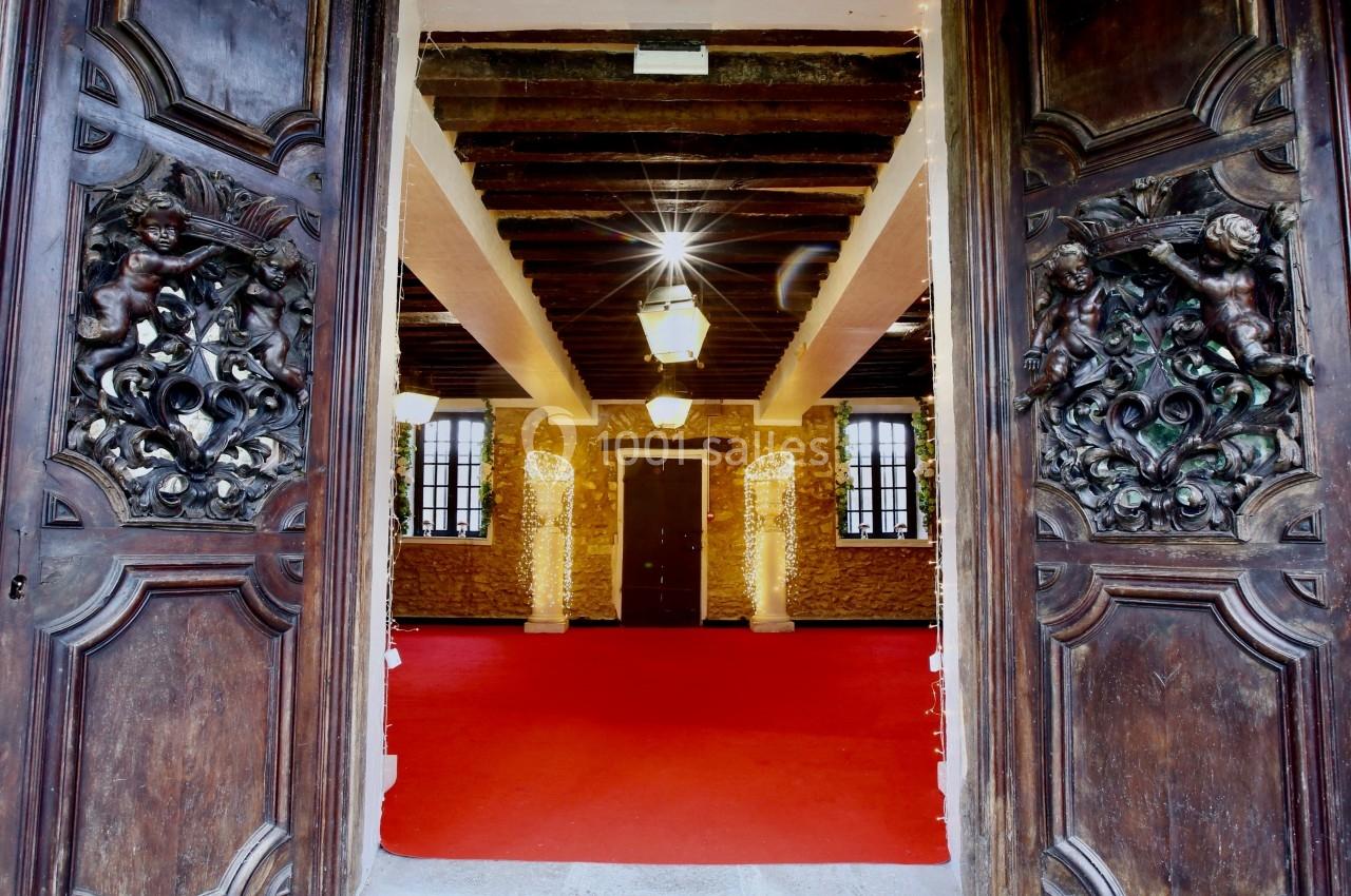 Portes en bois sculpté ouvertes sur une salle avec un sol rouge, des murs en pierre et des poutres apparentes.