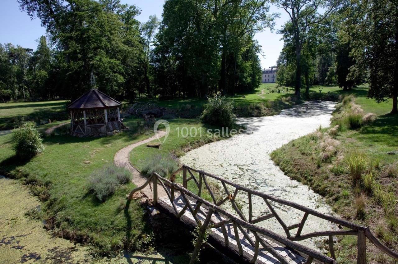 Un pont en bois traverse un ruisseau bordé de végétation dans un parc arboré avec un pavillon et un bâtiment au loin.