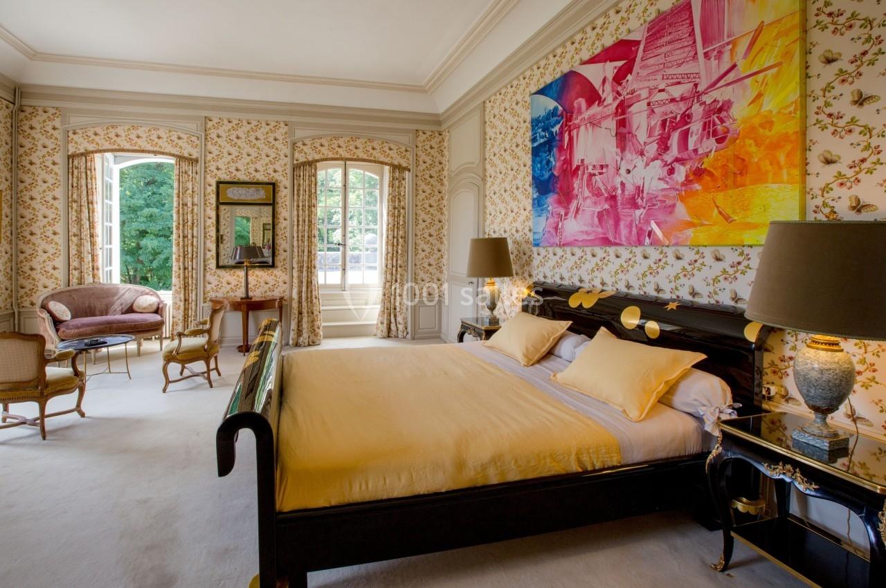 Chambre élégante avec lit double, murs décorés de motifs floraux, grande fenêtre et fauteuils dans un coin salon.
