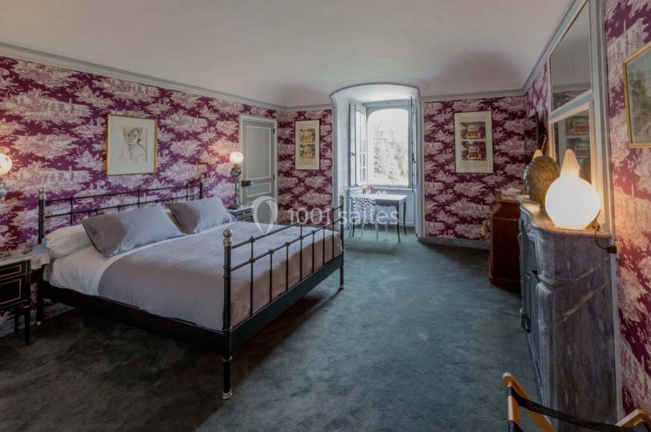 Chambre avec lit double, murs tapissés de motifs violets, fenêtre lumineuse et mobilier classique.