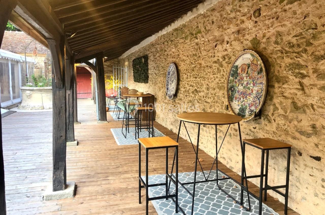 Terrasse en bois couverte avec tables hautes et tabourets, adossée à un mur en pierre décoré de mosaïques.