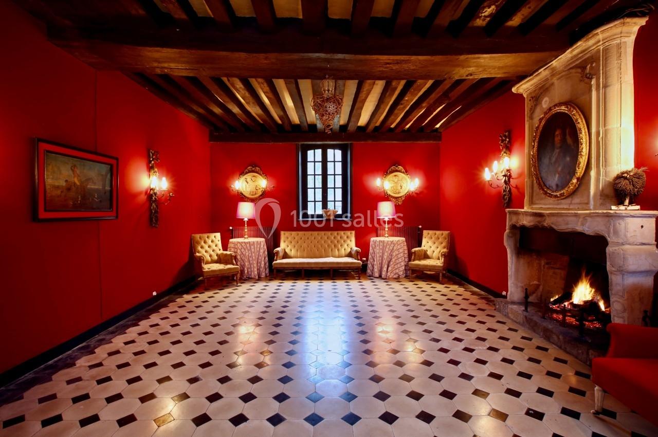 Salle avec murs rouges, cheminée allumée, plafond à poutres apparentes, mobilier classique et sol en damier.