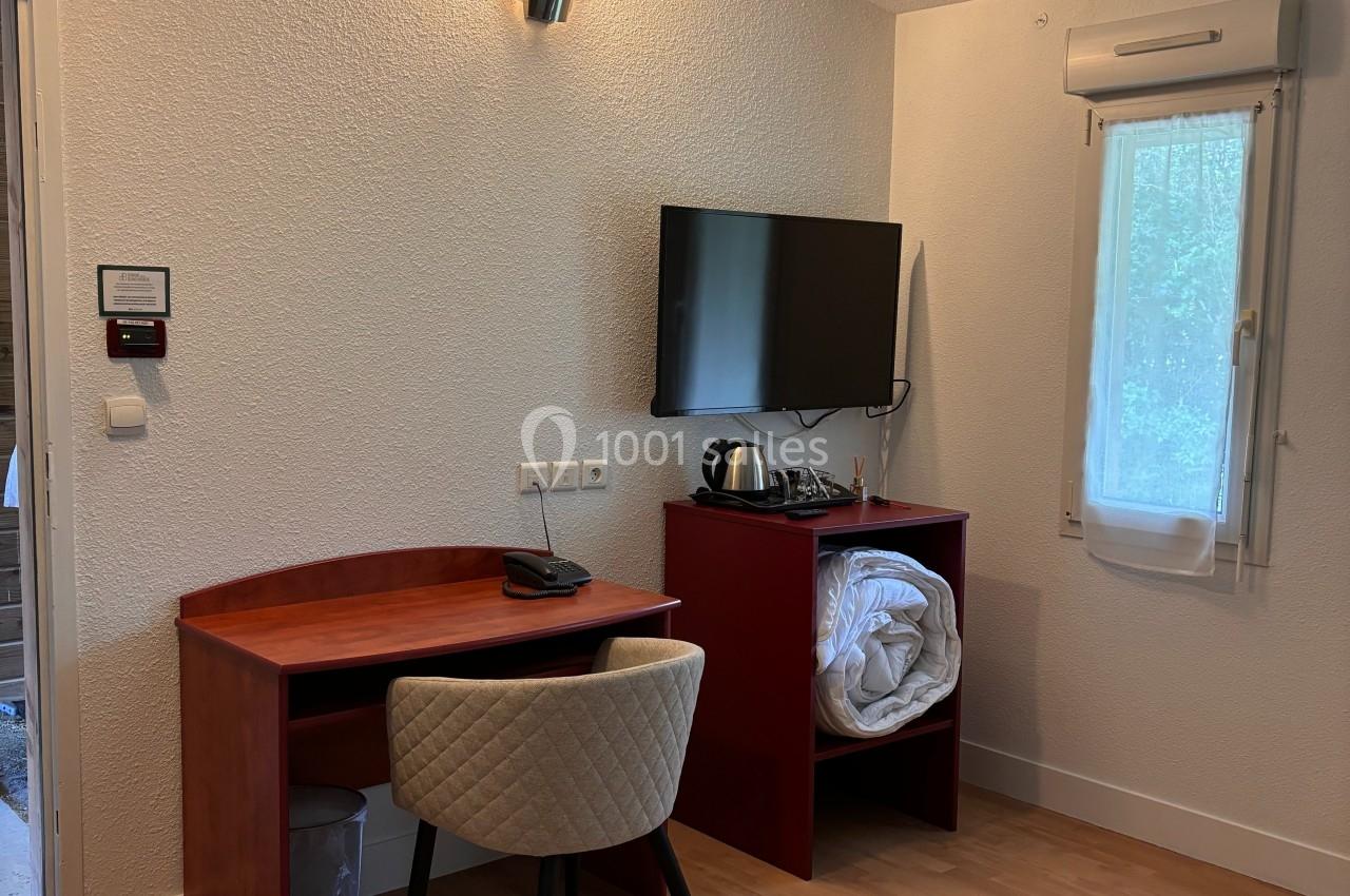 Bureau en bois avec chaise, télévision murale, étagère contenant une bouilloire et du linge, dans une pièce lumineuse.