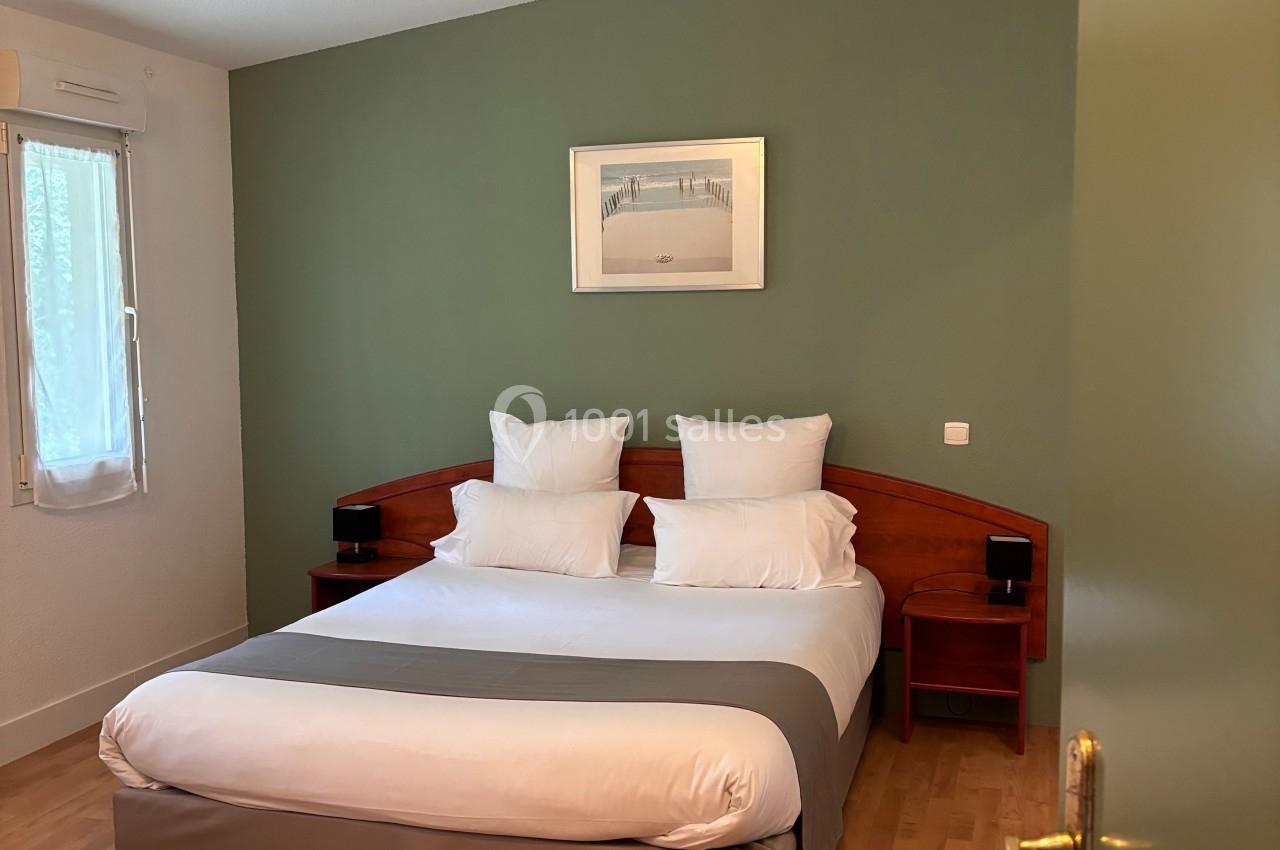 Chambre avec lit double, draps blancs, mur vert, parquet clair, fenêtre avec rideaux et cadre décoratif au mur.