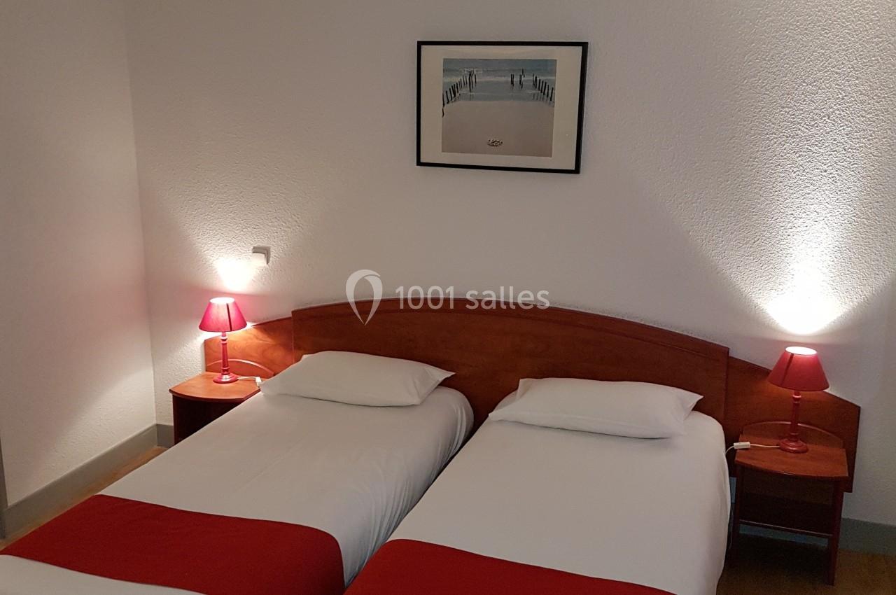 Deux lits simples avec draps blancs et chemins de lit rouges, encadrés par des lampes de chevet dans une chambre sobre.