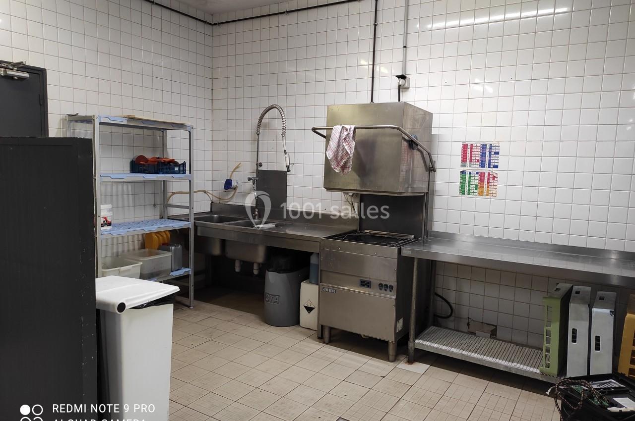 Cuisine professionnelle avec évier, étagères de rangement, lave-vaisselle industriel et murs carrelés blancs.