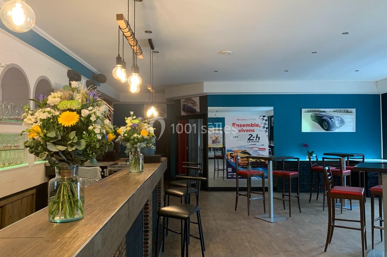 Intérieur d'un café moderne avec bar en bois, tabourets, tables hautes et bouquets de fleurs colorées.