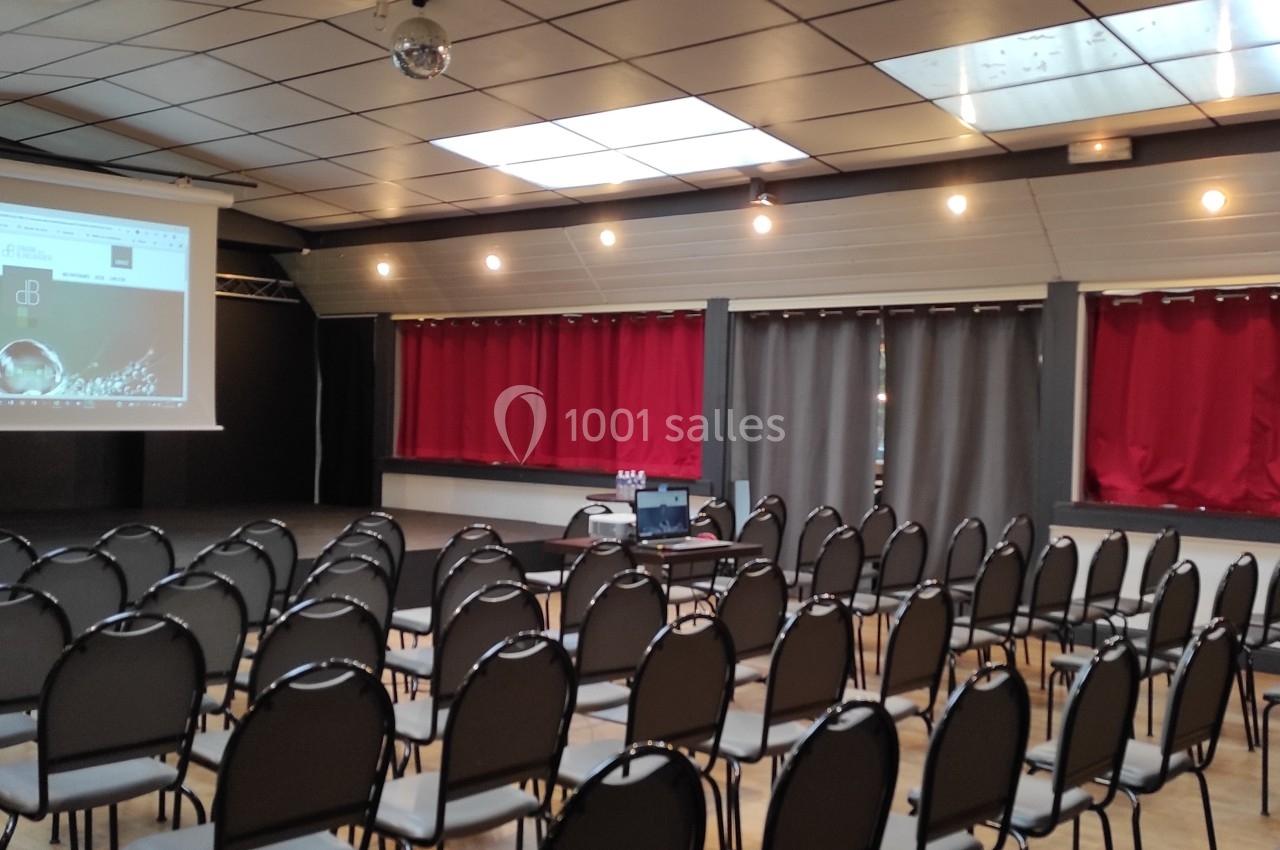 Salle de conférence avec des rangées de chaises vides, un écran de projection allumé et des rideaux rouges en arrière-plan.