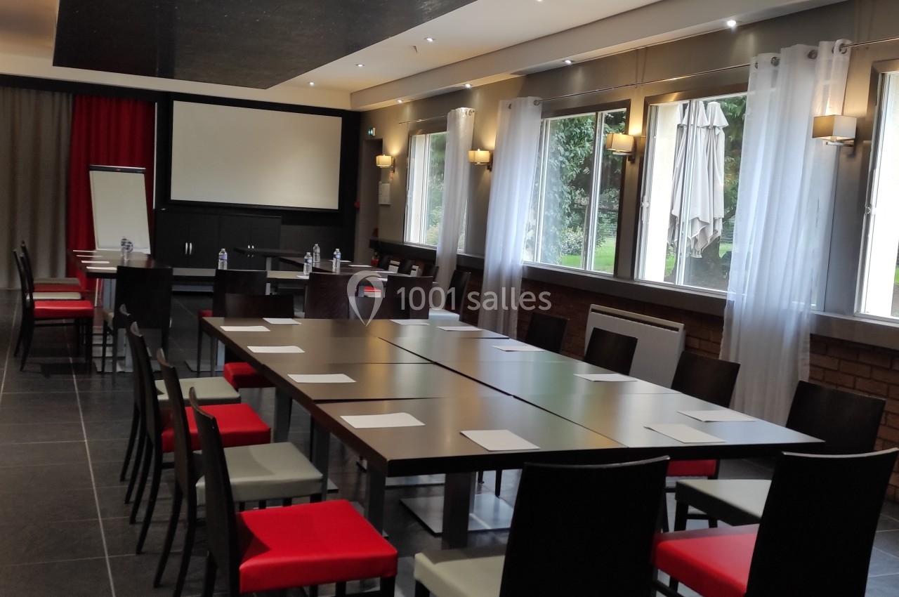Salle de réunion lumineuse avec une grande table rectangulaire, chaises rouges et noires, écran de projection et fenêtres…