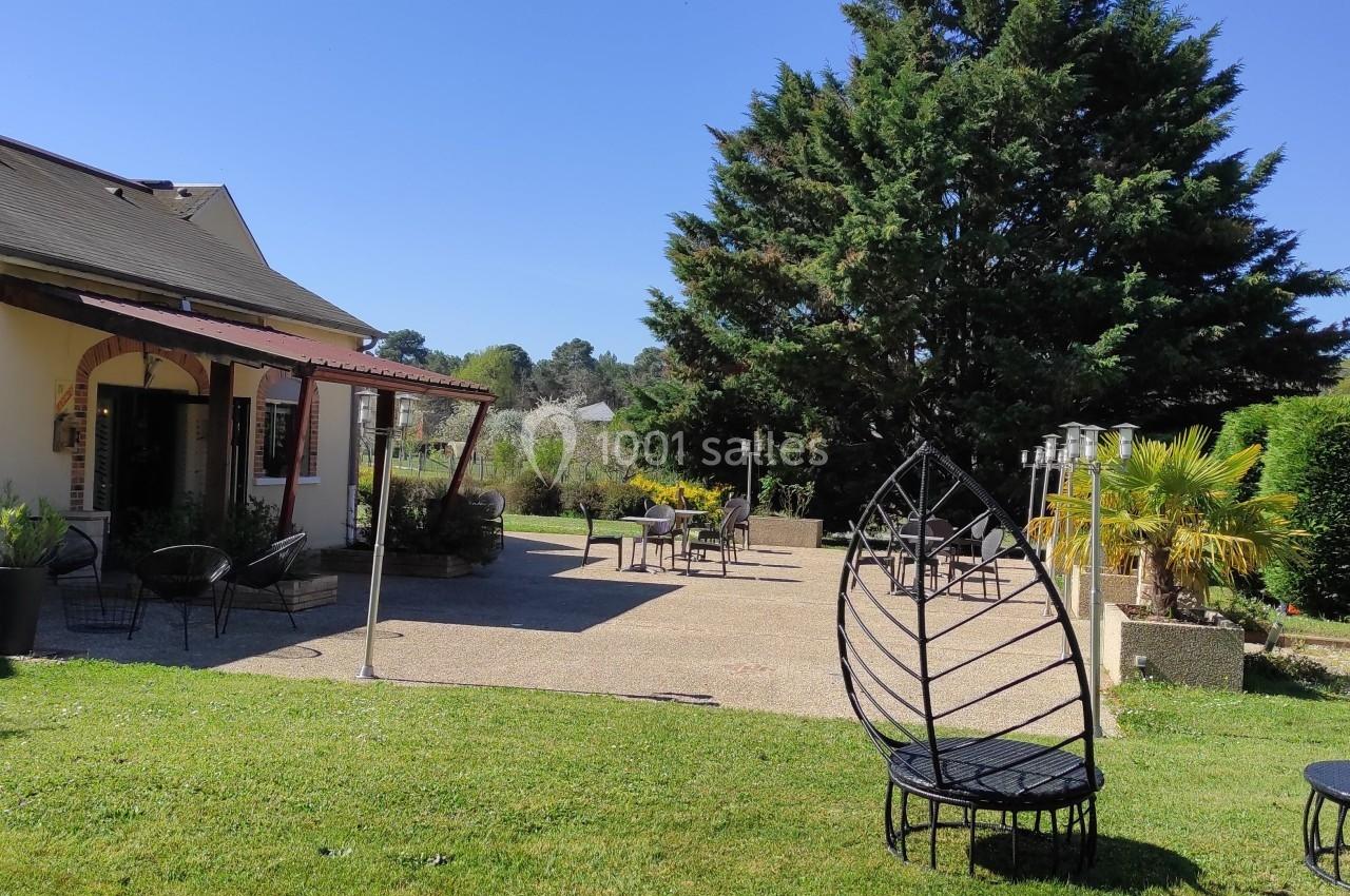 Terrasse d'une maison avec mobilier de jardin, pelouse, arbres et ciel dégagé en arrière-plan.