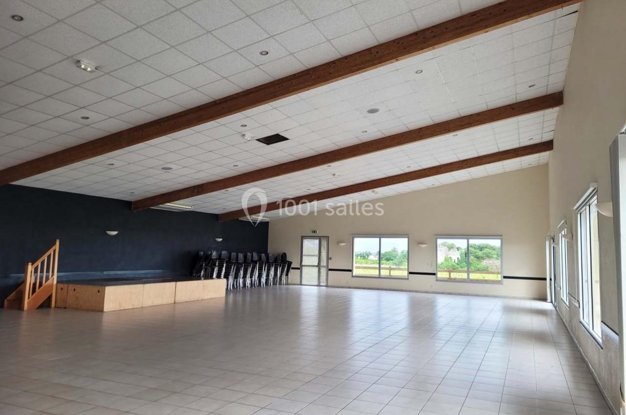 Salle spacieuse avec sol carrelé, scène en bois, chaises empilées et grandes fenêtres donnant sur un paysage verdoyant.