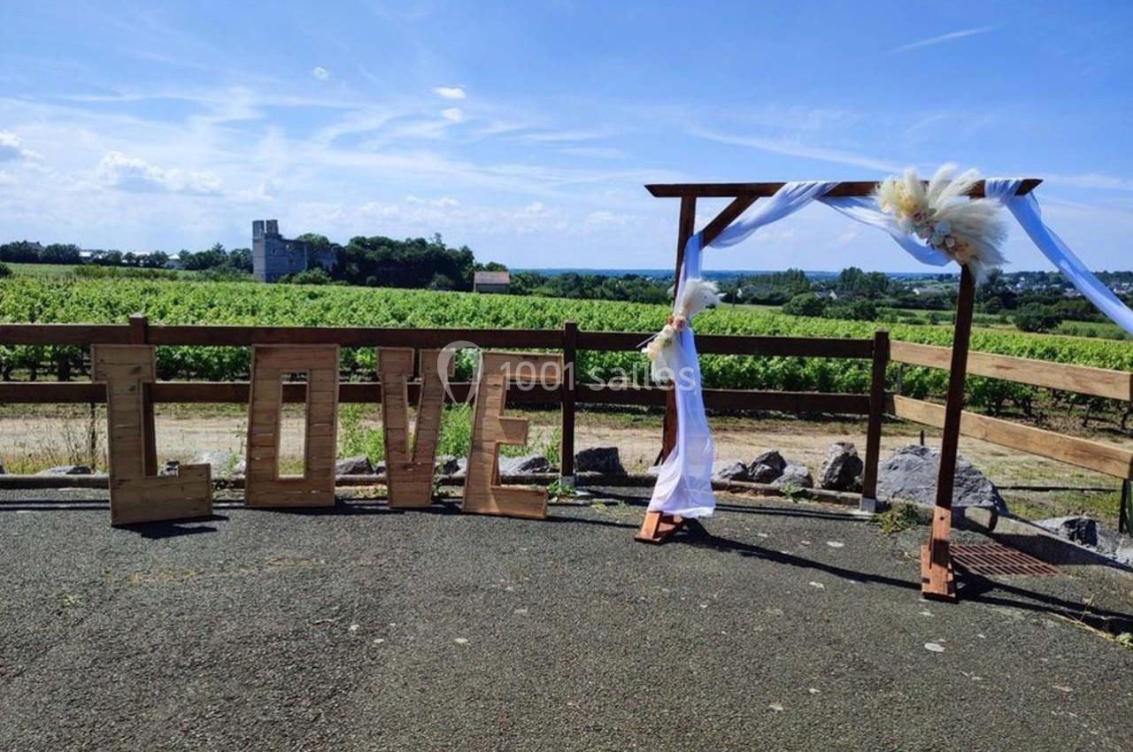 Structure en bois formant le mot ’LOVE’ et arche décorée de tissu blanc, devant un vignoble sous un ciel dégagé.