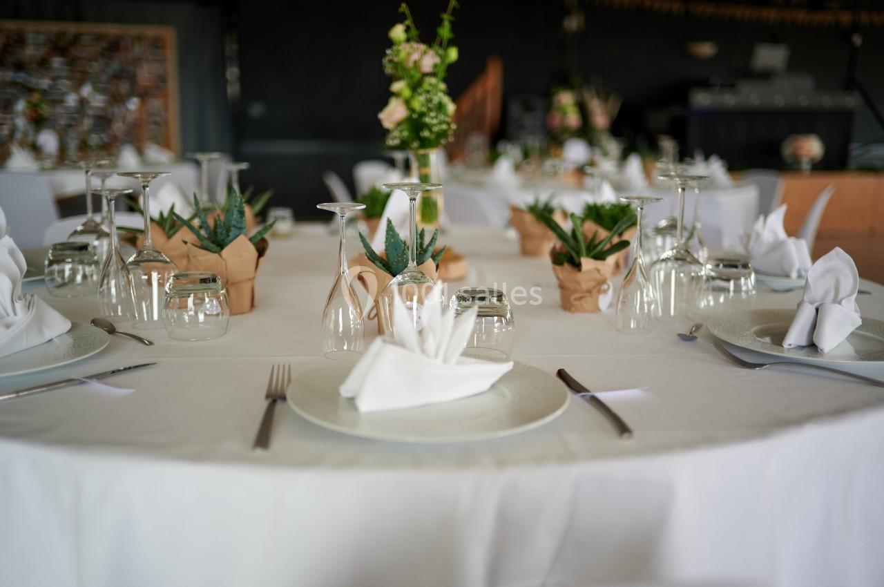 Table ronde dressée avec nappes blanches, verres, serviettes pliées et décorations florales au centre.