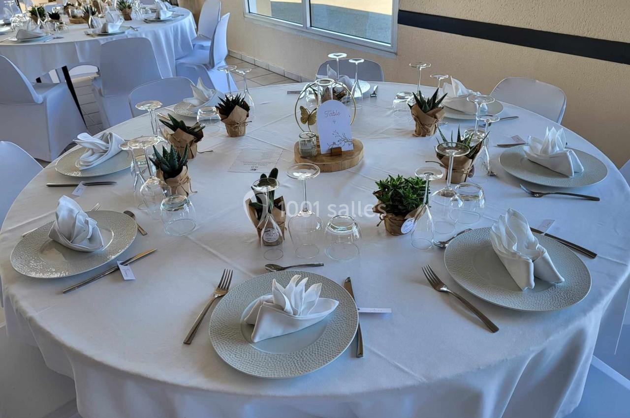 Table ronde dressée pour un repas, avec nappes blanches, serviettes pliées, couverts, verres et décorations végétales.