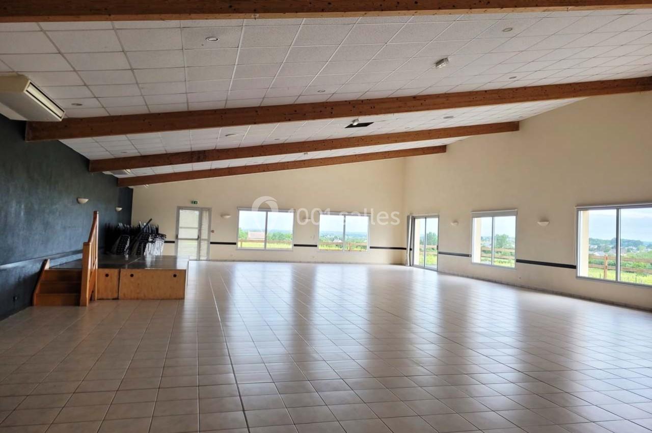 Salle spacieuse avec carrelage clair, poutres apparentes et grandes fenêtres offrant une vue sur l'extérieur.