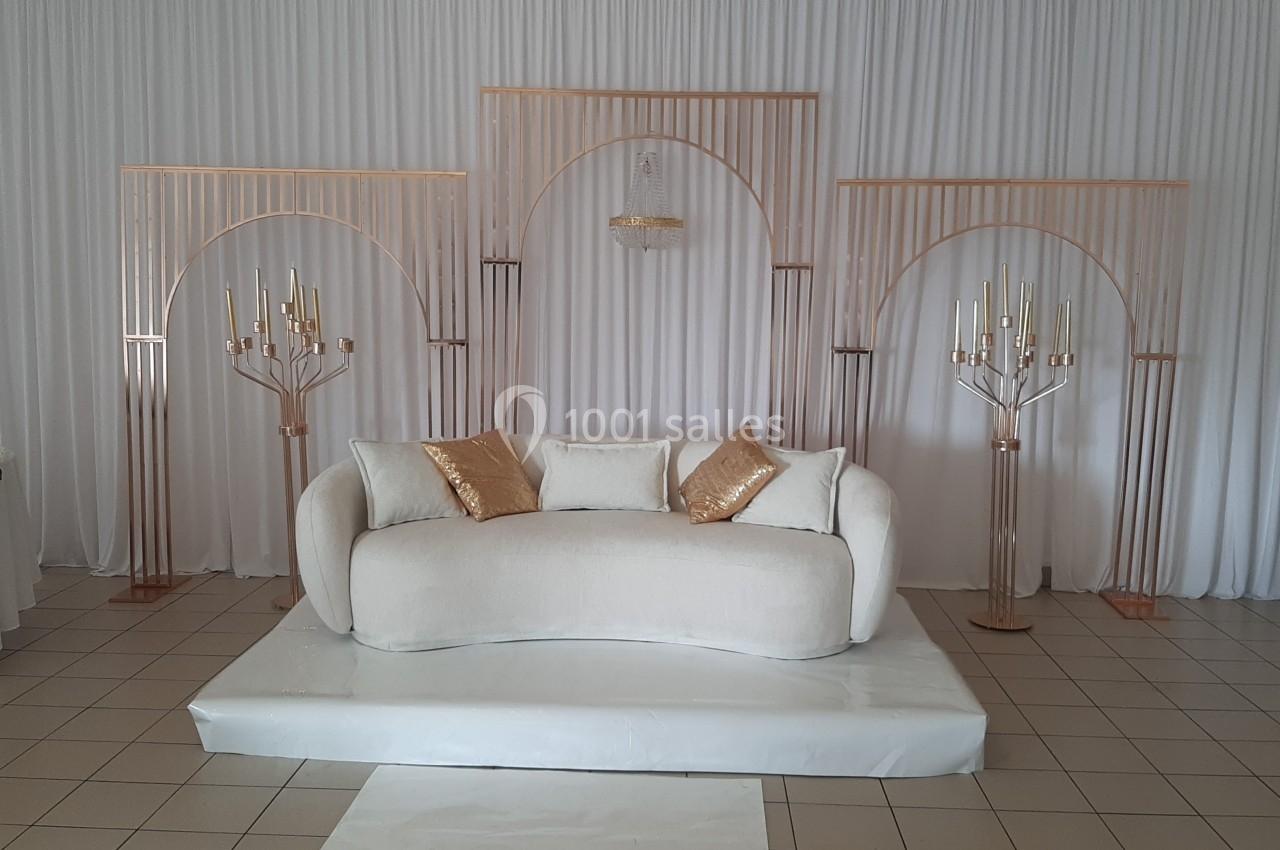 Canapé blanc avec coussins dorés, posé sur une estrade, entouré de structures décoratives dorées devant un rideau blanc.