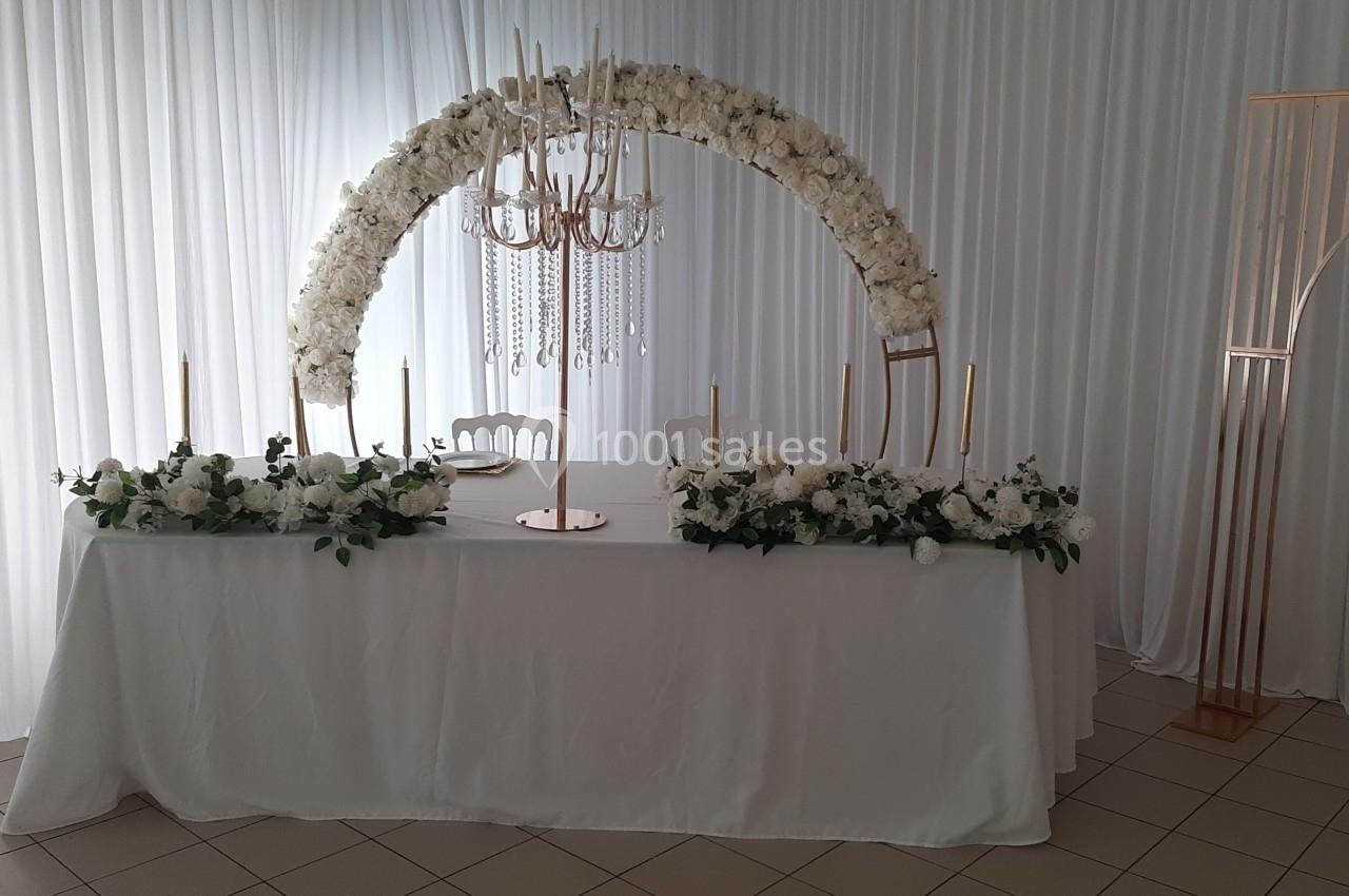 Table décorée pour un mariage avec nappes blanches, chandeliers dorés, fleurs blanches et arche florale en arrière-plan.